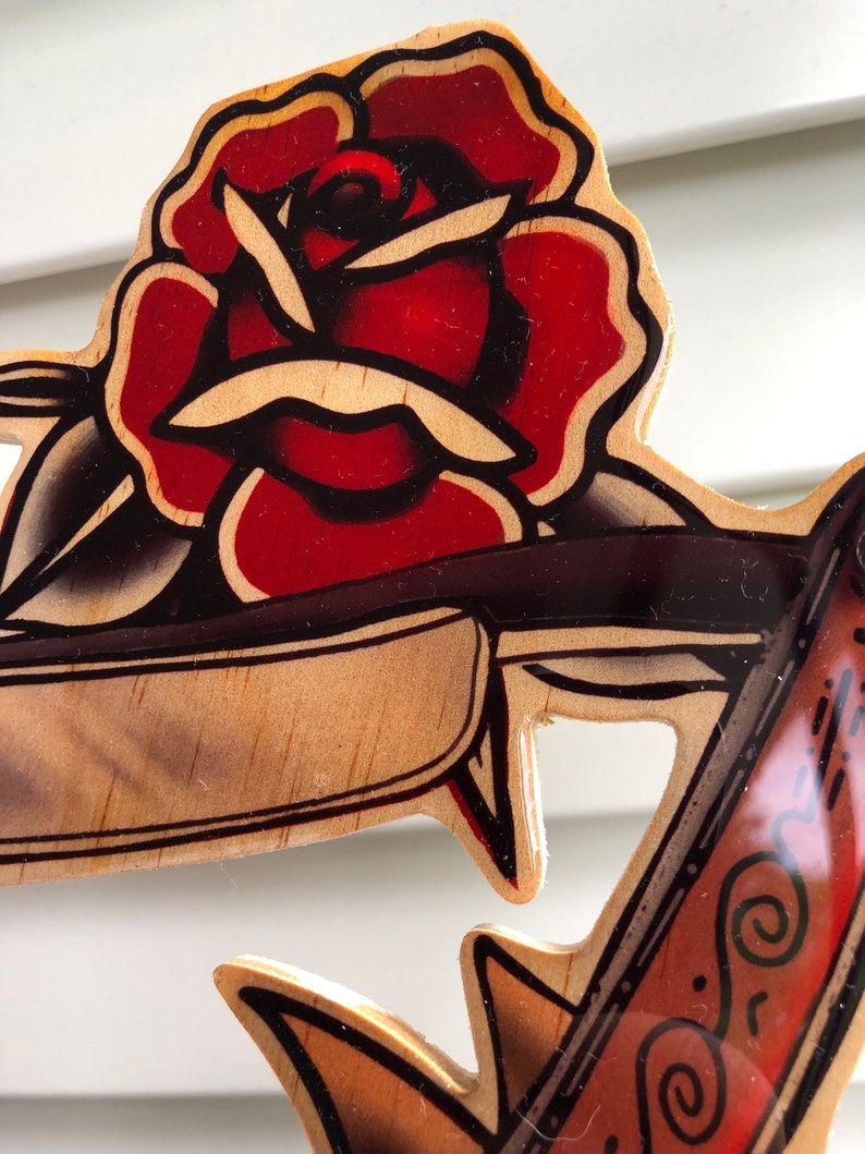May include: A wooden wall art piece with a red rose and an anchor. The rose is painted in a traditional tattoo style with black outlines and red and white shading. The anchor is painted in a similar style with brown, black, and white shading.