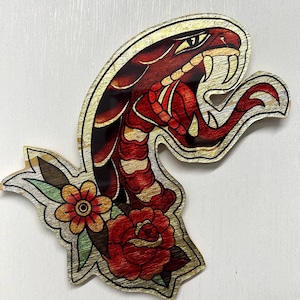 May include: Wooden art piece depicting a red snake with gold detailing and a floral arrangement. The snake's head is raised, and the design includes a red rose and other flowers. The artwork is in a vintage tattoo style.