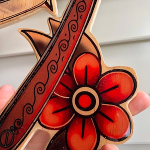May include: A wooden cutout of a red flower with a black outline and a red and black razor blade with a decorative pattern. The cutout is painted with a glossy finish.