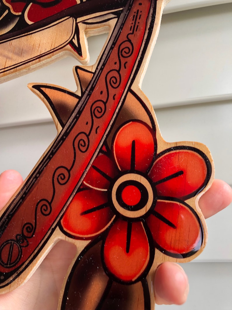 May include: A wooden cutout of a red flower with a black outline and a red and black razor blade with a decorative pattern. The cutout is painted with a glossy finish.