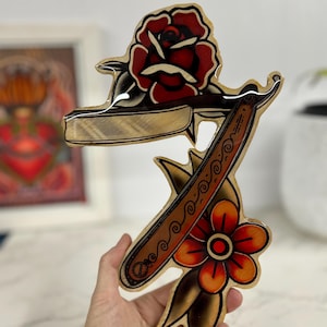 May include: A wooden wall art piece with a vintage tattoo style design featuring a straight razor, a rose, and a flower. The design is painted in red, orange, yellow, brown, and black.