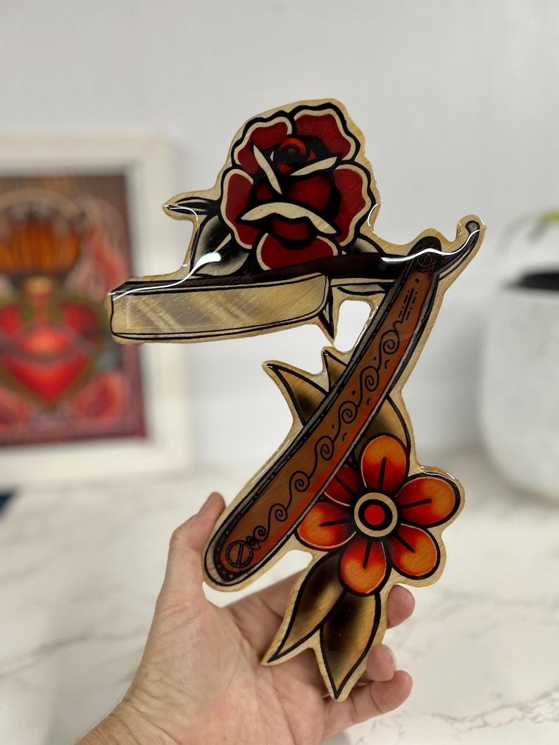 May include: A wooden wall art piece with a vintage tattoo style design featuring a straight razor, a rose, and a flower. The design is painted in red, orange, yellow, brown, and black.
