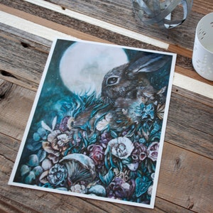 May include: A watercolor painting of a rabbit in a forest setting. The rabbit is brown and white and is surrounded by lush green foliage and flowers. The moon is visible in the background, shining a soft light on the scene.