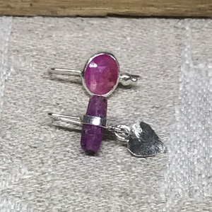 May include: A pair of silver earrings with pink gemstone accents. One earring features a round gemstone, while the other has a long, rectangular gemstone. The earrings have a heart-shaped charm.