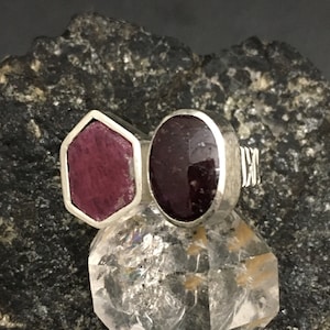 Pode incluir: Um anel de prata com duas pedras preciosas vermelhas engastadas em forma hexagonal e oval. O anel está sobre um cristal de quartzo transparente.