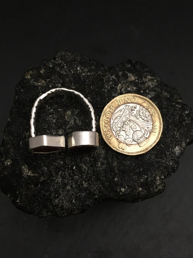 May include: A silver ring with two red gemstone settings. The ring is shown next to a British one pound coin for size comparison.