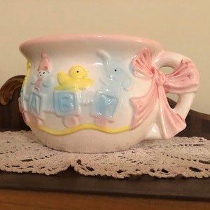 May include: A white ceramic potty with a pink bow and a blue, yellow, and pink cartoon design. The design features a duck, a bear, and a train.