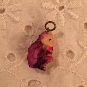 May include: A small, purple and pink plastic charm of a monkey holding a red apple. The charm has a gold loop for attaching to a necklace or bracelet.