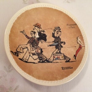 May include: A decorative plate with a beige background and a cream-colored rim. The plate features a cartoon illustration of people walking, with text that reads "Promenade à Paris" and "Vote for Al". Signed "Norman Rockwell".