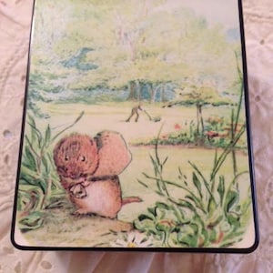 May include: A wooden music box with a hand-painted image of a small brown rabbit in a green meadow. The rabbit is holding a small object in its paws. The background features a green meadow with trees and a small figure in the distance.