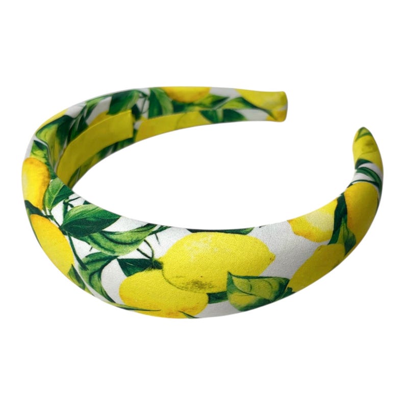 May include: A padded headband featuring a vibrant lemon pattern. The headband is primarily white, with bright yellow lemons and green leaves scattered across the surface. The interior is a solid yellow.