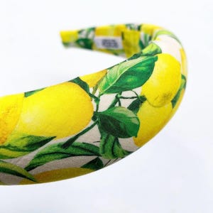 May include: A padded headband featuring a vibrant lemon print. The design showcases bright yellow lemons and green leaves against a cream-colored background. The headband is a stylish accessory for hair styling.