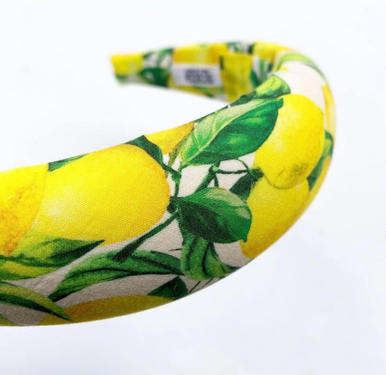 May include: A padded headband featuring a vibrant lemon print. The design showcases bright yellow lemons and green leaves against a cream-colored background. The headband is a stylish accessory for hair styling.
