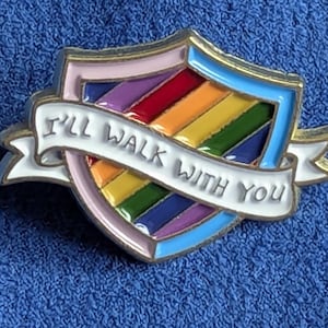 Può includere: Una spilla smaltata colorata a forma di scudo con un design arcobaleno. La spilla ha uno striscione bianco con la scritta "I'll Walk With You". La spilla ha un bordo rosa, blu e oro.