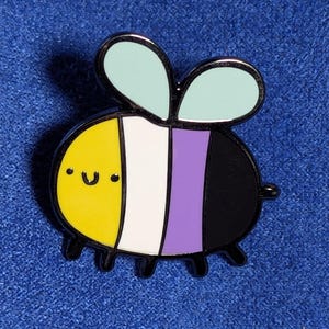 Puede incluir: Un pin de abeja con la bandera del orgullo no binario, con cuerpo amarillo, franja blanca, franja morada y cuerpo negro. La abeja tiene una cara sonriente y alas celestes. El pin está sobre un fondo azul.