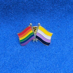 Puede incluir: Un pin dorado con dos banderas cruzadas. Una bandera muestra los colores del arcoíris de la bandera del orgullo LGBTQ+, y la otra bandera tiene rayas moradas, blancas y negras. El pin está sobre un fondo azul.