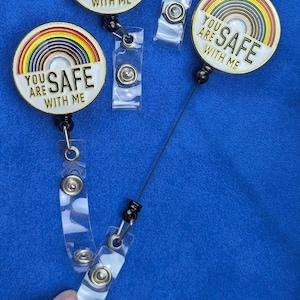 Op de afbeelding: Vier badge reels met een regenboogontwerp en de tekst "YOU ARE SAFE WITH ME" op een witte achtergrond. De reels hebben een intrekbaar koord en een doorzichtige plastic clip. De achtergrond is effen blauw.