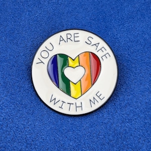 Può includere: Spilla rotonda in smalto bianco con un design a cuore arcobaleno. Il cuore è diviso in strisce verticali rosse, arancioni, gialle, verdi, blu e viola. Il testo "YOU ARE SAFE WITH ME" è scritto in nero attorno al cuore.