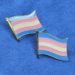 Op de afbeelding: Twee emaille pins met het ontwerp van de transgender pride vlag. Elke pin heeft een gouden rand en een vlag met horizontale strepen in lichtblauw, roze en wit. De pins worden getoond op een blauwe stoffen achtergrond.