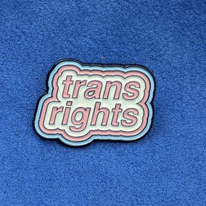 May include: A pin with the words "trans rights" in a white, pink, and blue design. The pin has a black border and is set against a blue background.