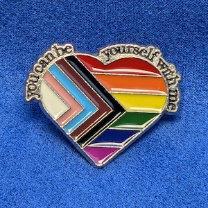 Peut inclure: Une épingle en forme de cœur avec un motif arc-en-ciel et l'inscription argentée "you can be yourself with me". L'épingle présente les couleurs du drapeau de la fierté LGBTQ+.