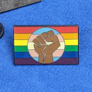 Peut inclure: Une broche émaillée rectangulaire présentant un fond de drapeau arc-en-ciel horizontal avec un emblème circulaire central contenant un poing levé marron. Le motif intègre des bandes rouges, orange, jaunes, vertes, bleues et violettes, avec un cercle rose clair, blanc et bleu clair.