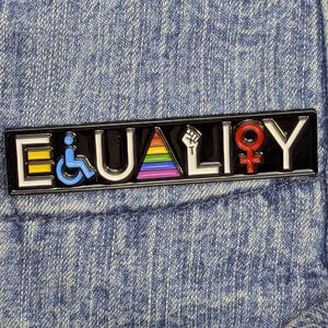Peut inclure: Épinglette en émail noir avec le mot "EQUALITY" en lettres blanches. L'épinglette présente des symboles pour le handicap, les LGBTQ+ et le féminisme. Le fond est une veste en jean bleue.