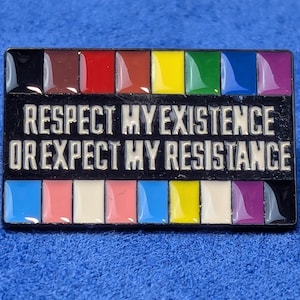 Może przedstawiać: Prostokątna przypinka z czarnym tłem i białym napisem "RESPECT MY EXISTENCE OR EXPECT MY RESISTANCE". Przypinka posiada tęczę kolorów u góry i na dole, w tym brązowy, czerwony, żółty, zielony, niebieski i fioletowy.