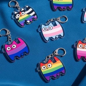 Puede incluir: Llaveros con forma de gato con diseños de banderas LGBTQ+ diferentes. Incluyen banderas del arcoíris, transgénero, bisexual y lesbiana. Los llaveros tienen anillos de metal plateado.