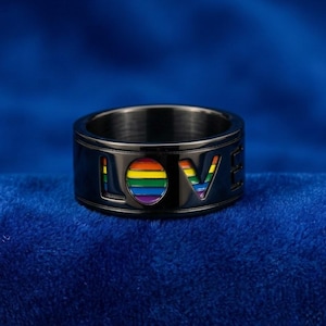 Peut inclure: Une bague noire avec le mot "LOVE" découpé, révélant un motif aux couleurs de l'arc-en-ciel. La bague est ornée des couleurs du drapeau de la fierté LGBTQ+ et se détache sur un fond bleu.