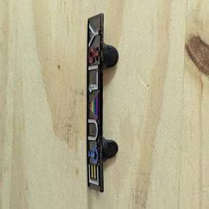 May include: A rectangular enamel pin with the word "YOUTH" in colorful letters. The pin has a black border and two black pin backs. The background is a light wood grain.