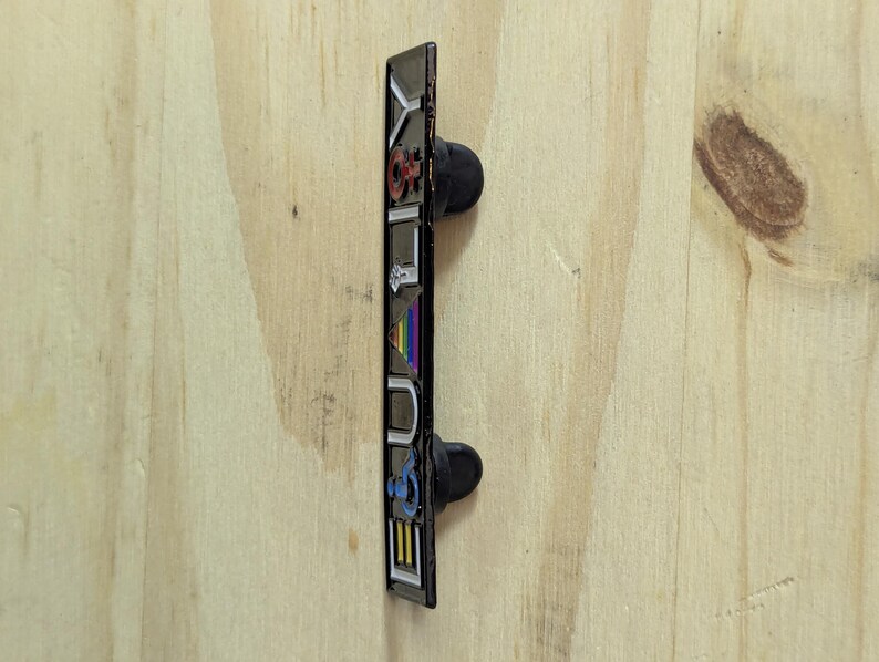 May include: A rectangular enamel pin with the word "YOUTH" in colorful letters. The pin has a black border and two black pin backs. The background is a light wood grain.