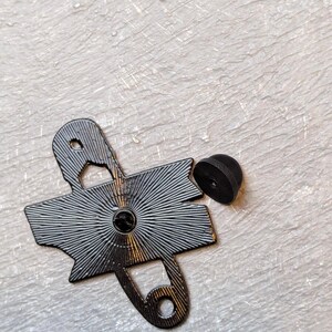 May include: A black enamel pin shaped like a safety pin, with a radiating design on the back. The pin has a small, round, black backing. The pin is on a textured, off-white surface.