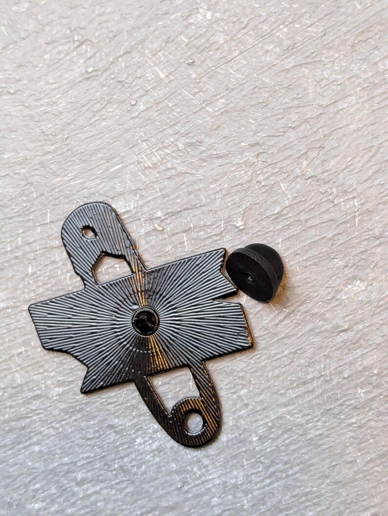 May include: A black enamel pin shaped like a safety pin, with a radiating design on the back. The pin has a small, round, black backing. The pin is on a textured, off-white surface.