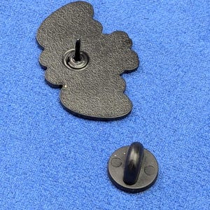 May include: A black enamel pin with a detailed design, featuring a pin back and a separate clutch. The pin has a textured surface and a rounded shape. The clutch is a small, round disc with a curved post.