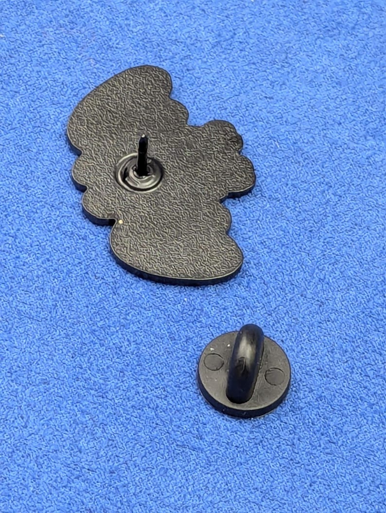 May include: A black enamel pin with a detailed design, featuring a pin back and a separate clutch. The pin has a textured surface and a rounded shape. The clutch is a small, round disc with a curved post.