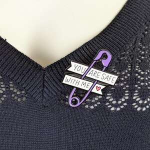 May include: A purple safety pin enamel pin with the text "YOU ARE SAFE WITH ME" on a white banner. The pin includes a small pink heart. The pin is attached to a dark blue sweater.
