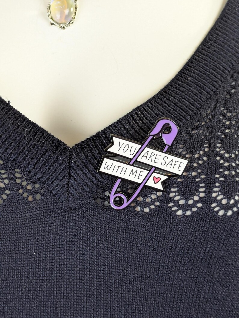 May include: A purple safety pin enamel pin with the text "YOU ARE SAFE WITH ME" on a white banner. The pin includes a small pink heart. The pin is attached to a dark blue sweater.