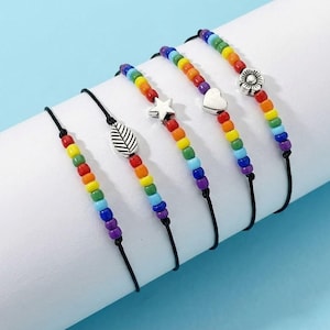 May include: Five beaded bracelets with rainbow-colored beads and black cords. Each bracelet features a different silver charm: a leaf, a star, a heart, and a flower. The bracelets are displayed on a white surface against a light blue background.