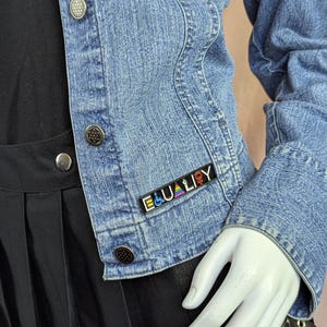 May include: A light blue denim jacket with silver buttons. The jacket has a small pin that reads "EQUALITY" in white letters with a rainbow design and gender symbols. The jacket is worn over a black top and skirt.