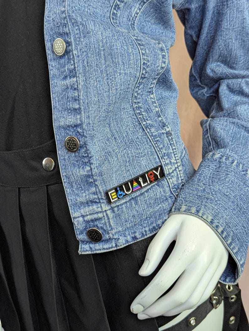 May include: A light blue denim jacket with silver buttons. The jacket has a small pin that reads "EQUALITY" in white letters with a rainbow design and gender symbols. The jacket is worn over a black top and skirt.