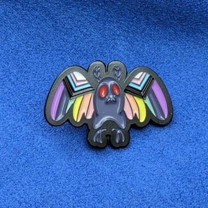 May include: A black enamel pin featuring a moth design with colorful wings. The moth has a dark gray body, red eyes, and wings with rainbow stripes. The pin is set against a blue background.