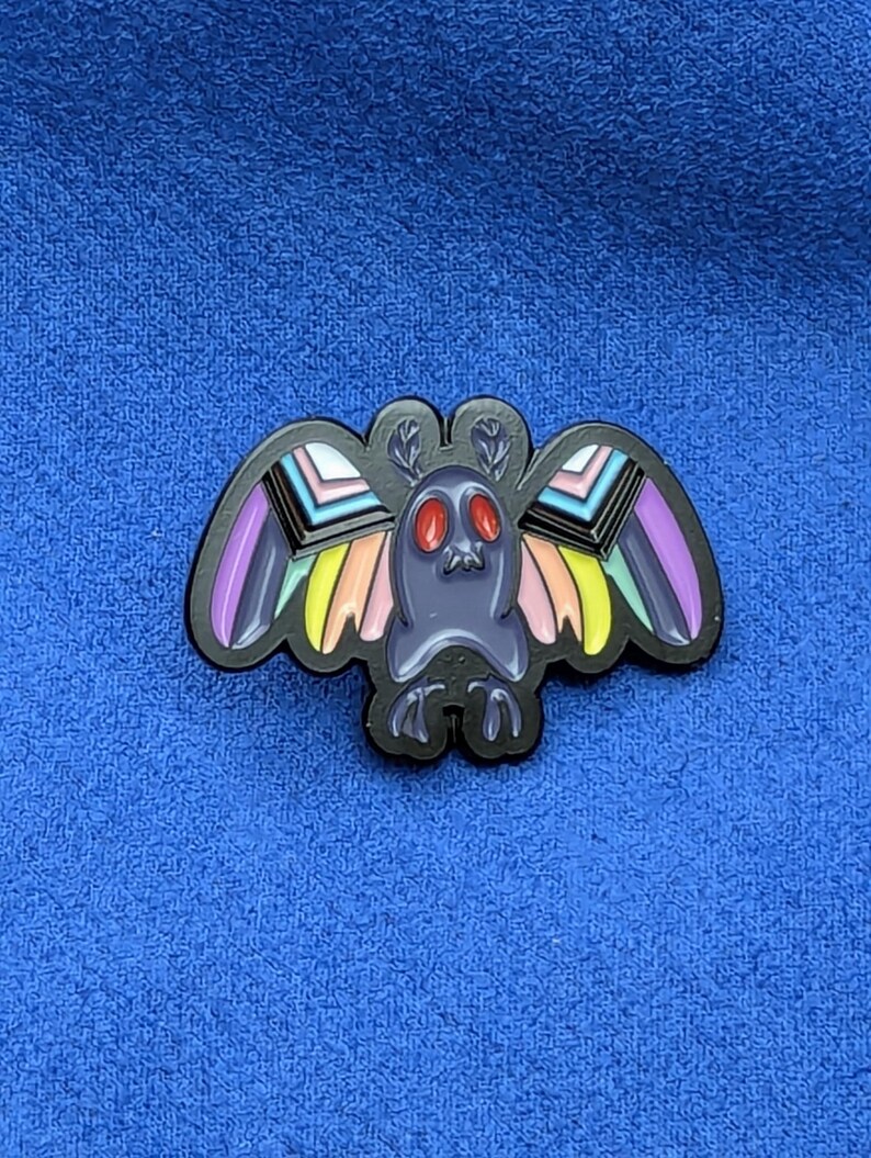 May include: A black enamel pin featuring a moth design with colorful wings. The moth has a dark gray body, red eyes, and wings with rainbow stripes. The pin is set against a blue background.