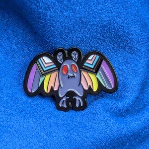 May include: A colorful enamel pin featuring a stylized moth design. The moth has a purple body, red eyes, and rainbow-colored wings with black outlines. The pin is set against a blue textured background.