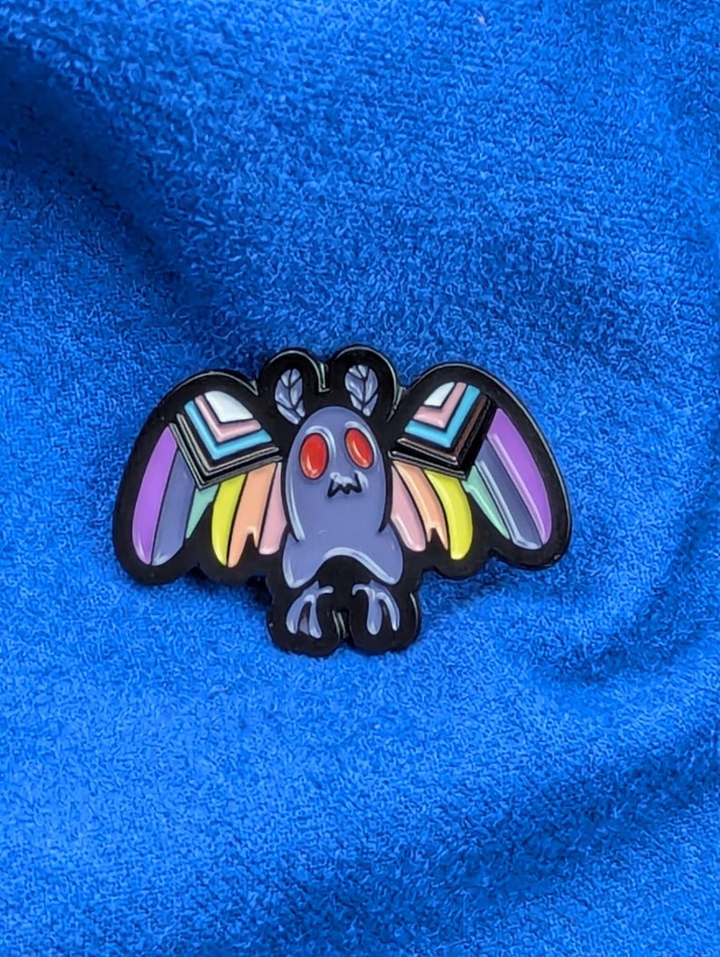 May include: A colorful enamel pin featuring a stylized moth design. The moth has a purple body, red eyes, and rainbow-colored wings with black outlines. The pin is set against a blue textured background.