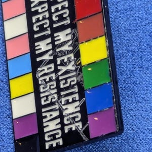 May include: A rectangular enamel pin with a black background and the words "RESPECT MY EXISTENCE" in white. The pin features a series of colorful stripes, including the transgender flag colors and a rainbow.