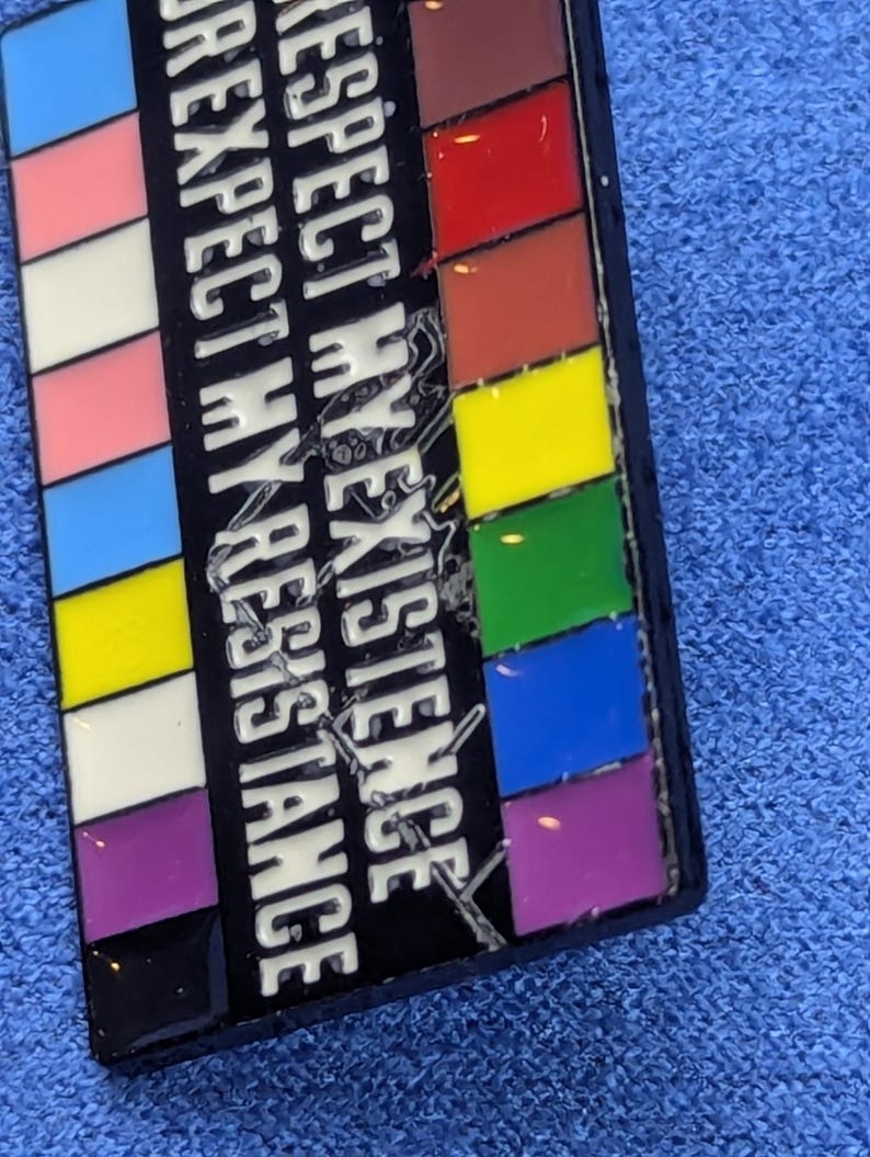 May include: A rectangular enamel pin with a black background and the words "RESPECT MY EXISTENCE" in white. The pin features a series of colorful stripes, including the transgender flag colors and a rainbow.