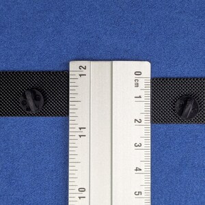 May include: Two black rectangular enamel pins with a textured surface, each with a black pin back, are displayed on a blue background. A ruler is placed between the pins to measure their size. The pins are approximately 1 inch in length.
