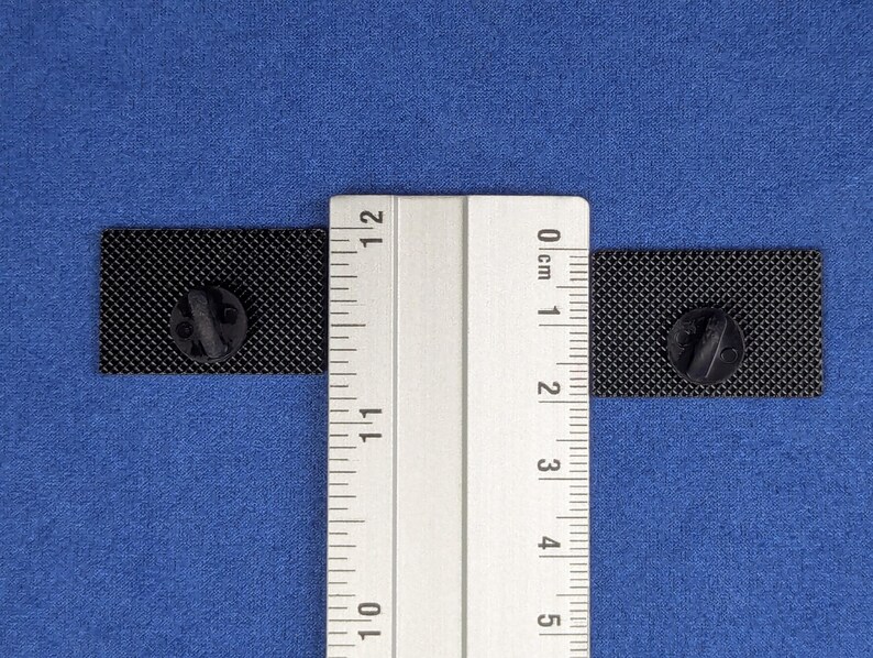 May include: Two black rectangular enamel pins with a textured surface, each with a black pin back, are displayed on a blue background. A ruler is placed between the pins to measure their size. The pins are approximately 1 inch in length.