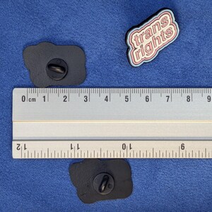 May include: Three enamel pins on a blue surface. One pin reads "trans rights" in pink and white lettering with a blue outline. Two black pins are also visible. A ruler is placed next to the pins.